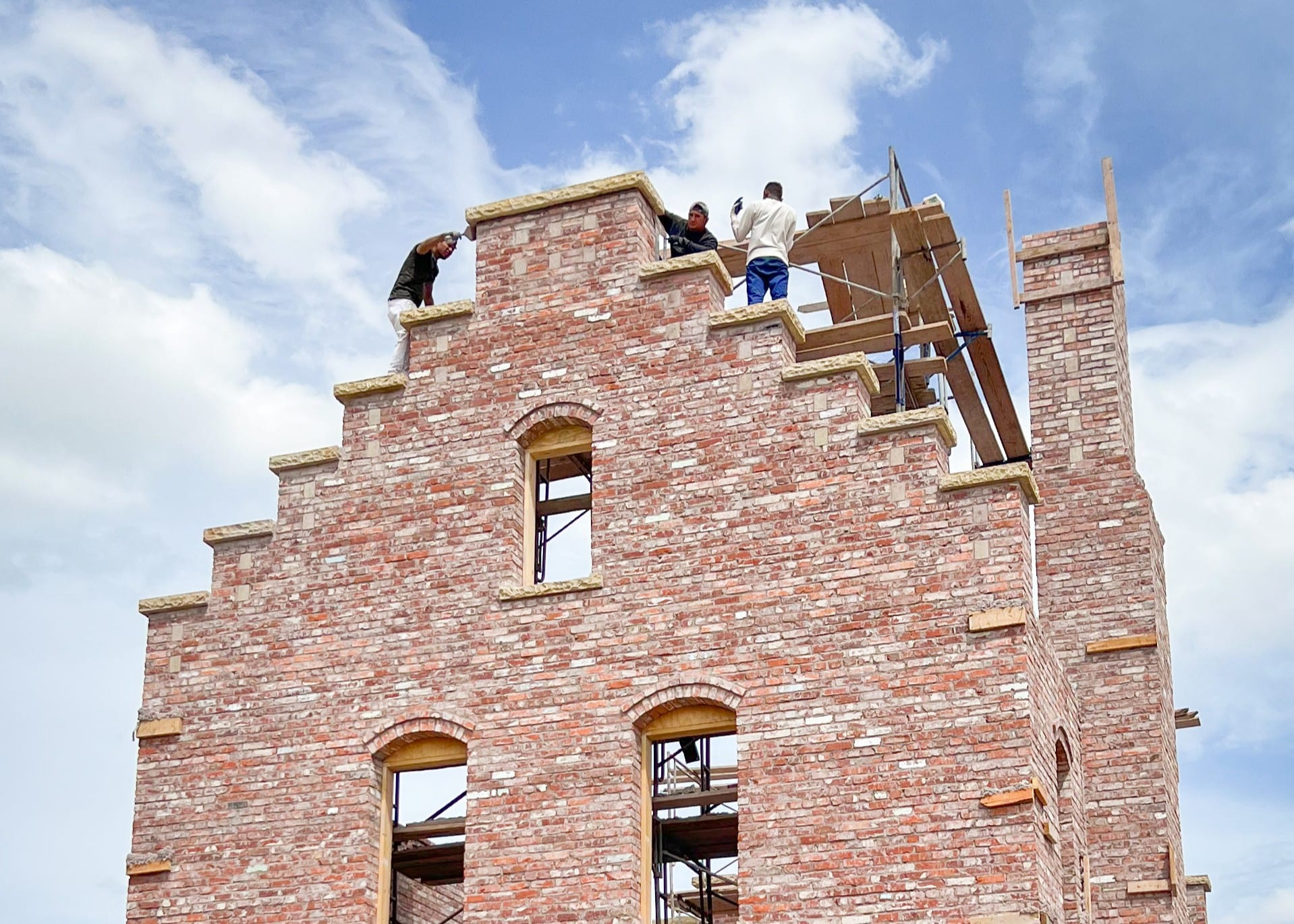 Masons laying the crow-stepped gable of a Building Culture structural brick tower under construction, against a bright sky.