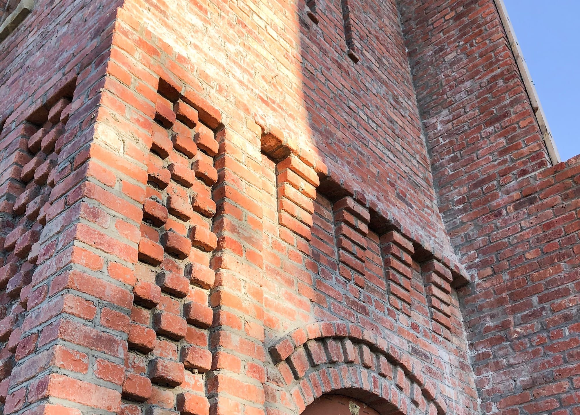 Detail of a Building Culture structural brick wall — corbeled brick patterning and an arched opening in warm raking light.