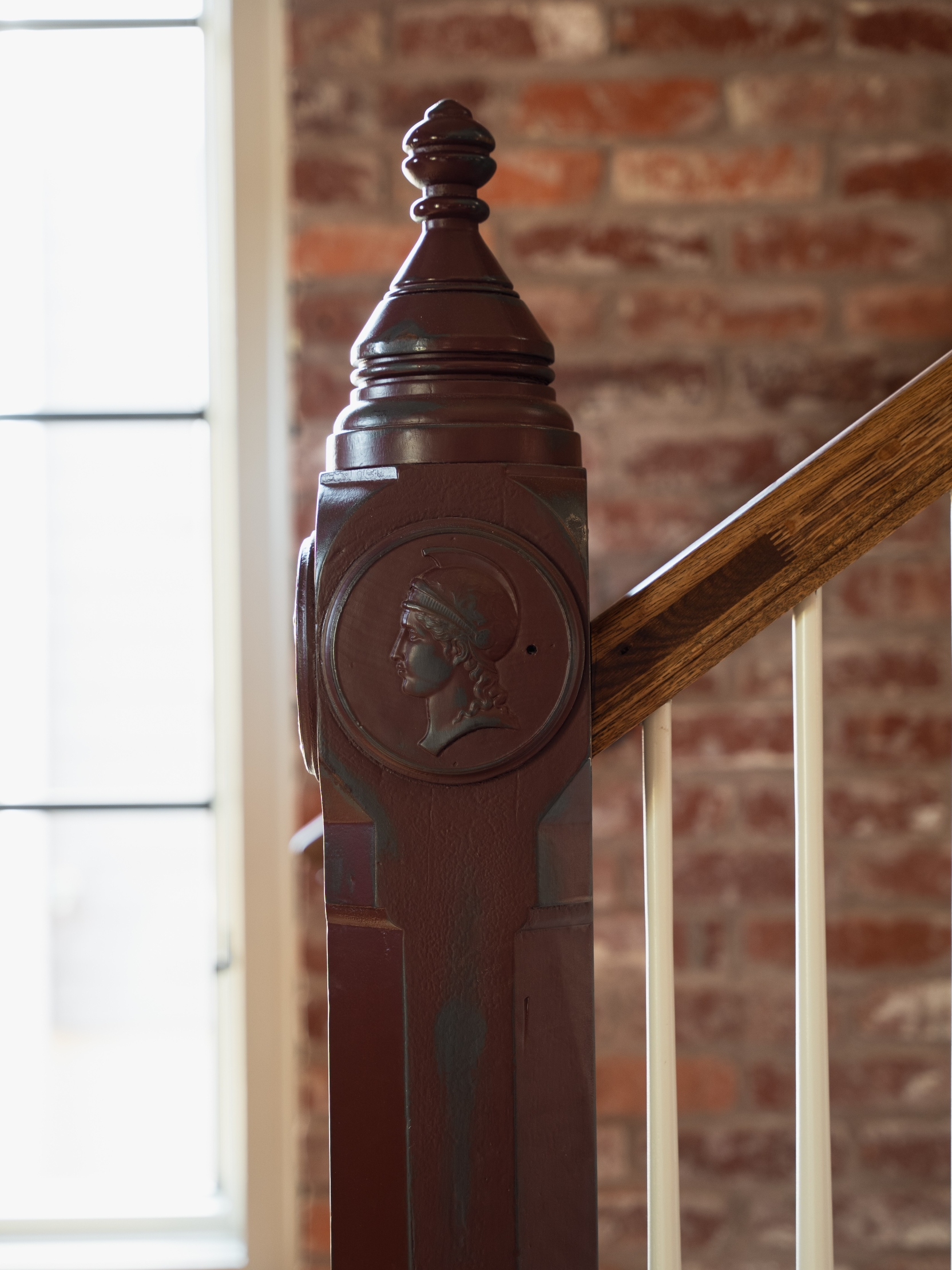 A handcrafted newel post with a classical female-profile medallion at the foot of an interior stair, brick wall behind.