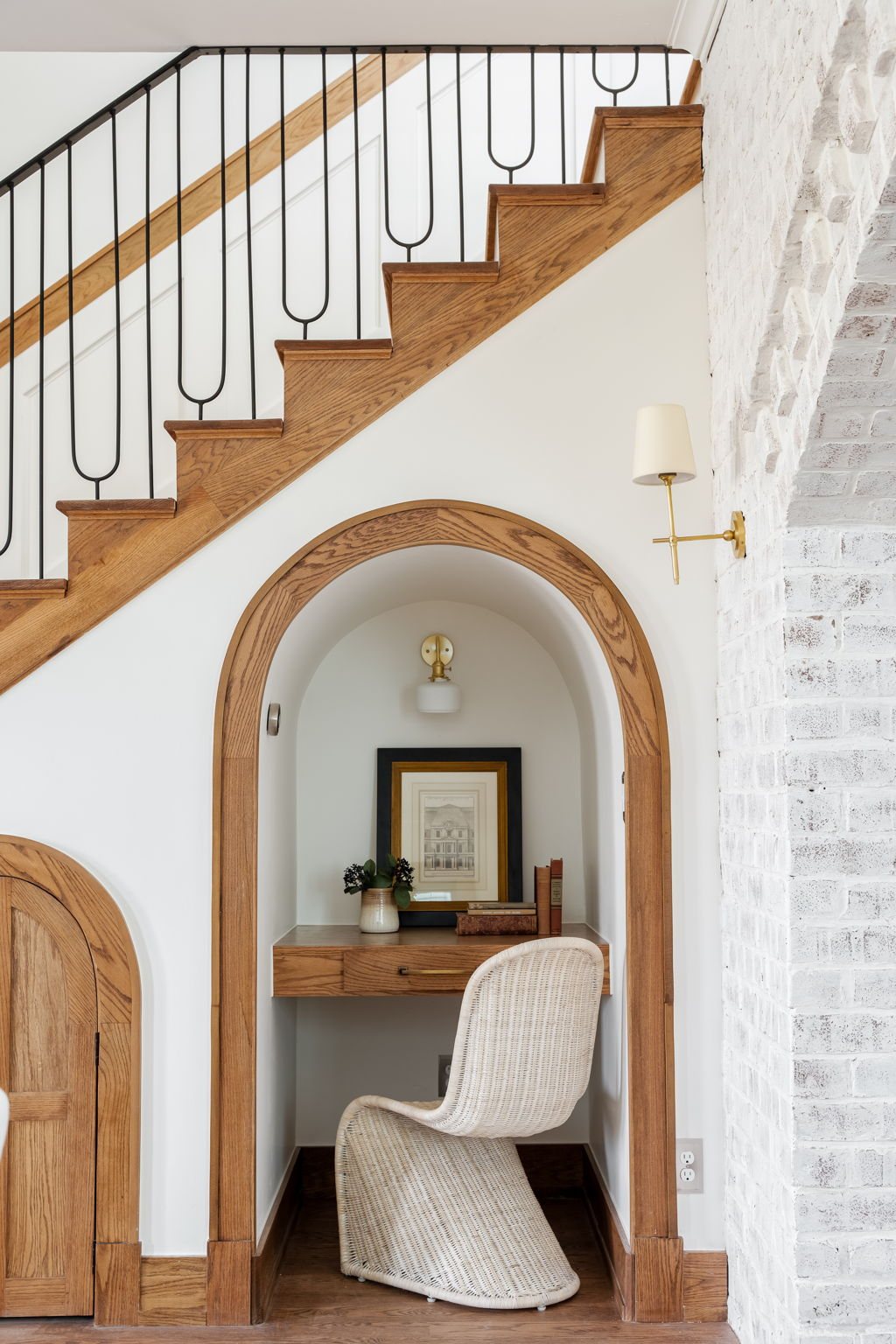 Built-in desk nook tucked beneath a staircase — oak millwork, arched threshold, whitewashed brick wall.