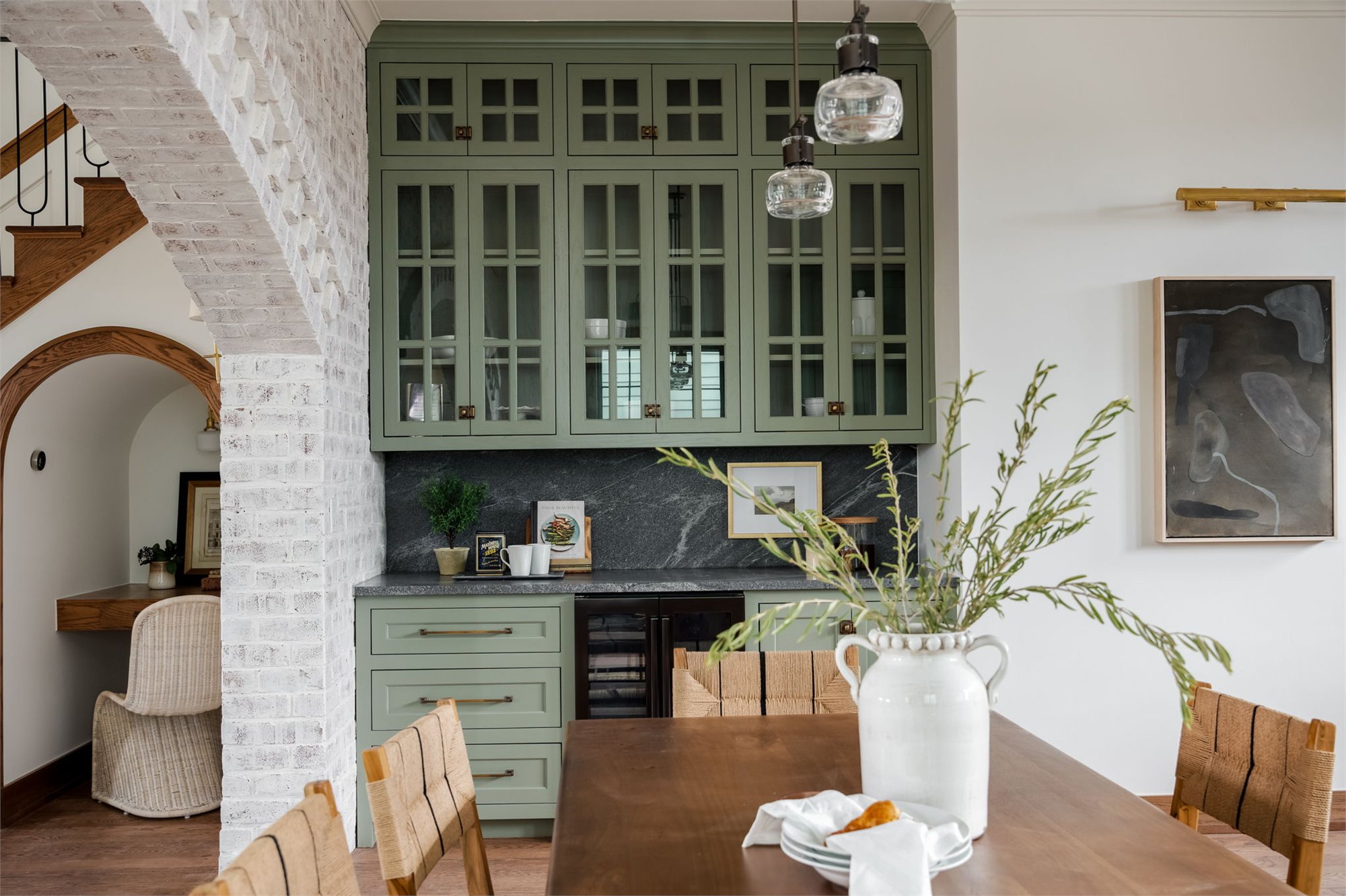 Butler's pantry with sage-green glass-front cabinetry, dark stone counter, and a brick archway opening into the next room.