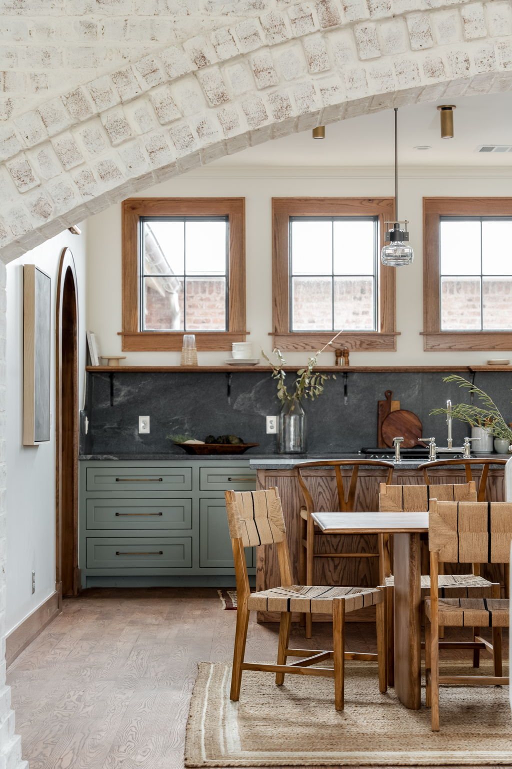 Kitchen under a whitewashed brick vaulted ceiling — oak windows, soapstone counters, sage cabinetry, woven dining chairs.