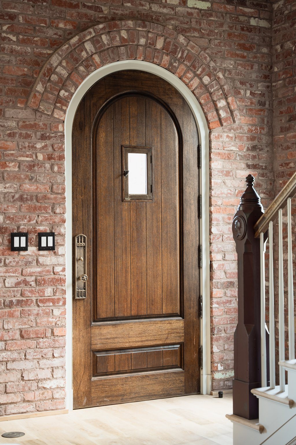 Arched walnut entry door set into a red-brick wall, with a stair newel and contrasting stone reveal.