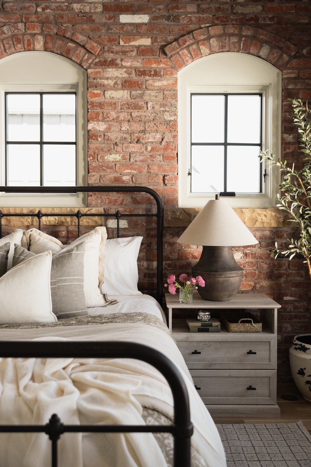 Bedroom with twin arched windows set into a red-brick wall, an iron bed frame, and layered linen bedding.