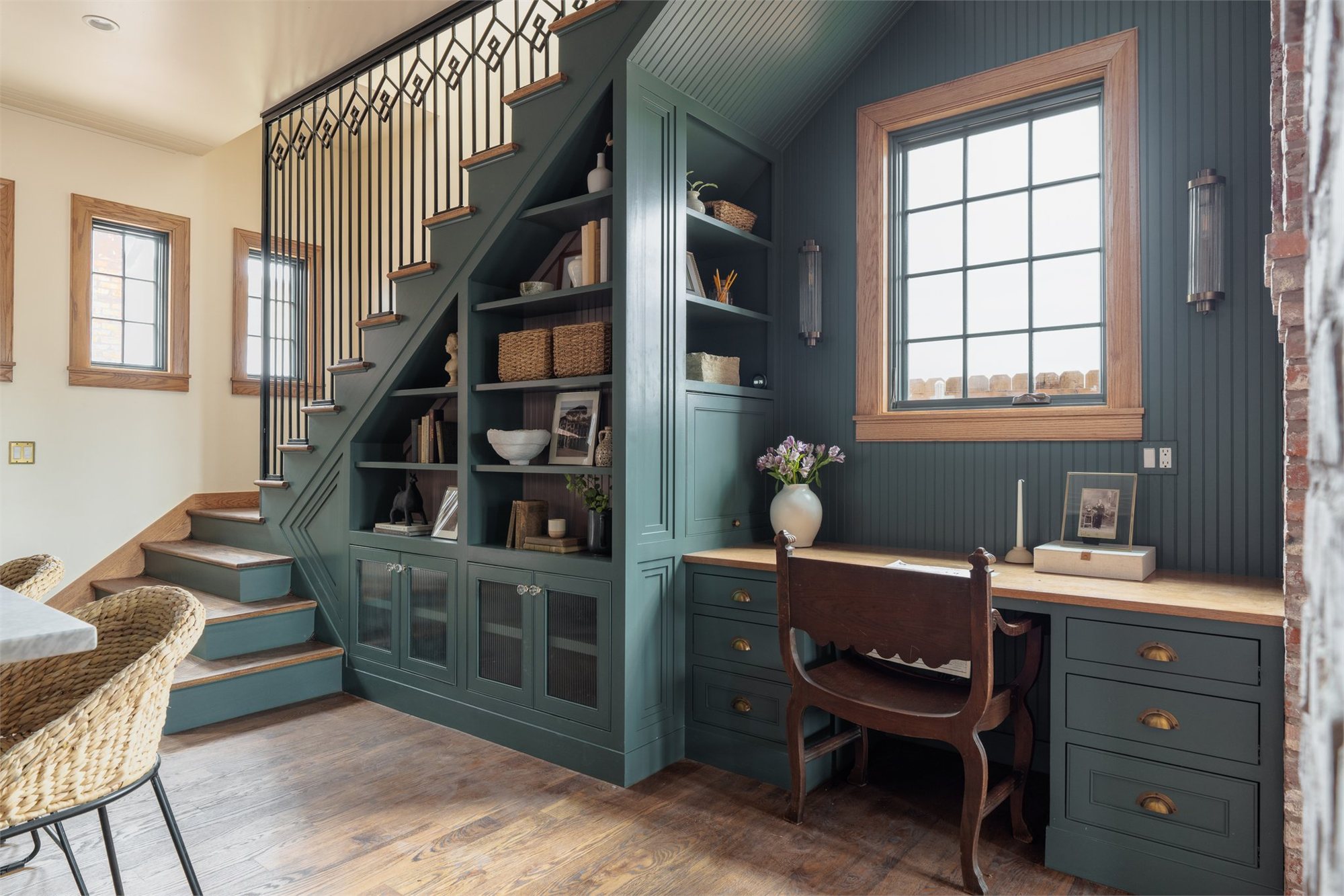 Dark teal millwork under an iron-baluster staircase — built-in bookcase, glass-front cabinets, and a desk on wide-plank floors.