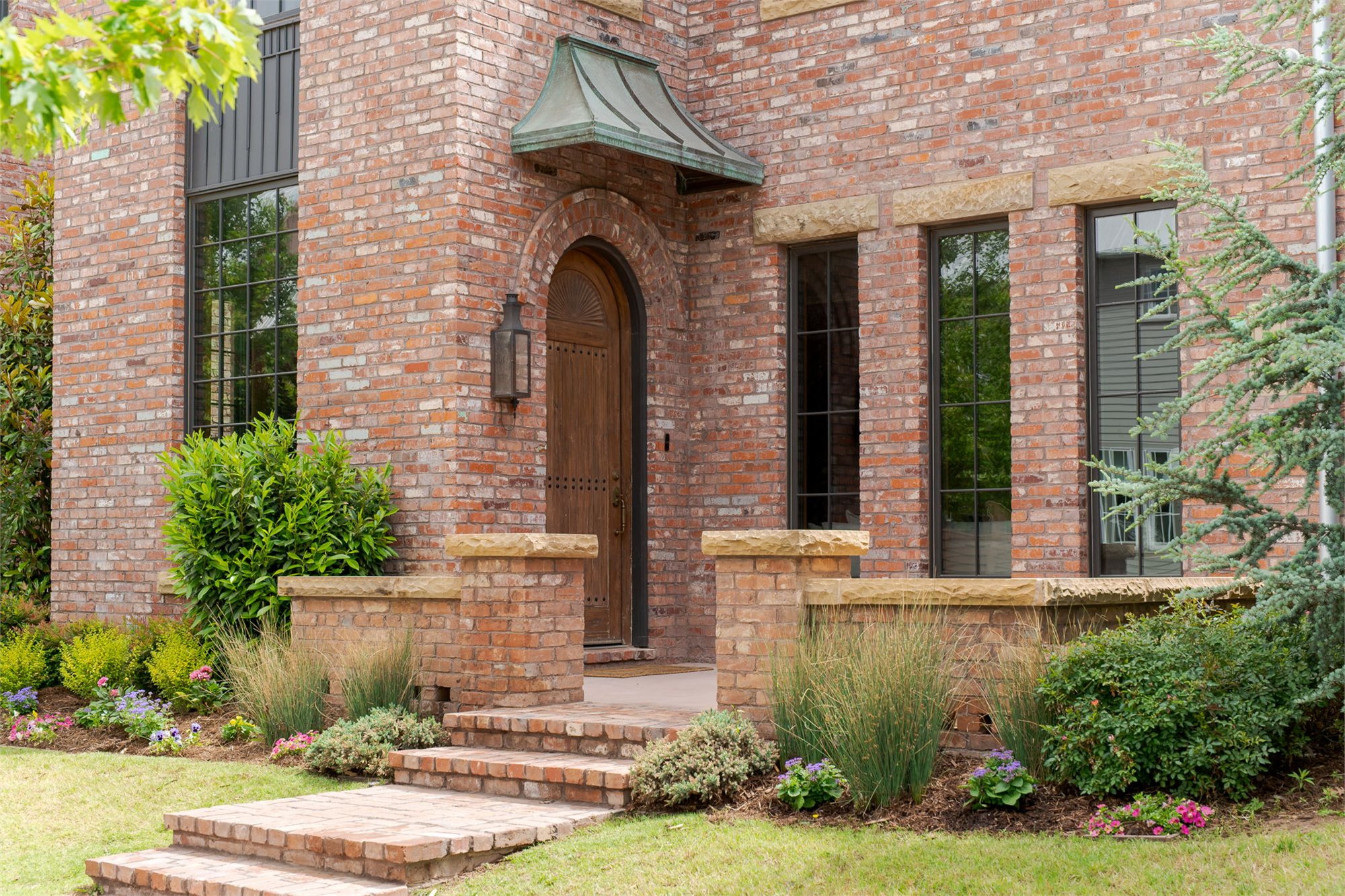 Arched walnut entry door under a copper hood, set in a red-brick facade with sandstone reveals and lavender plantings.