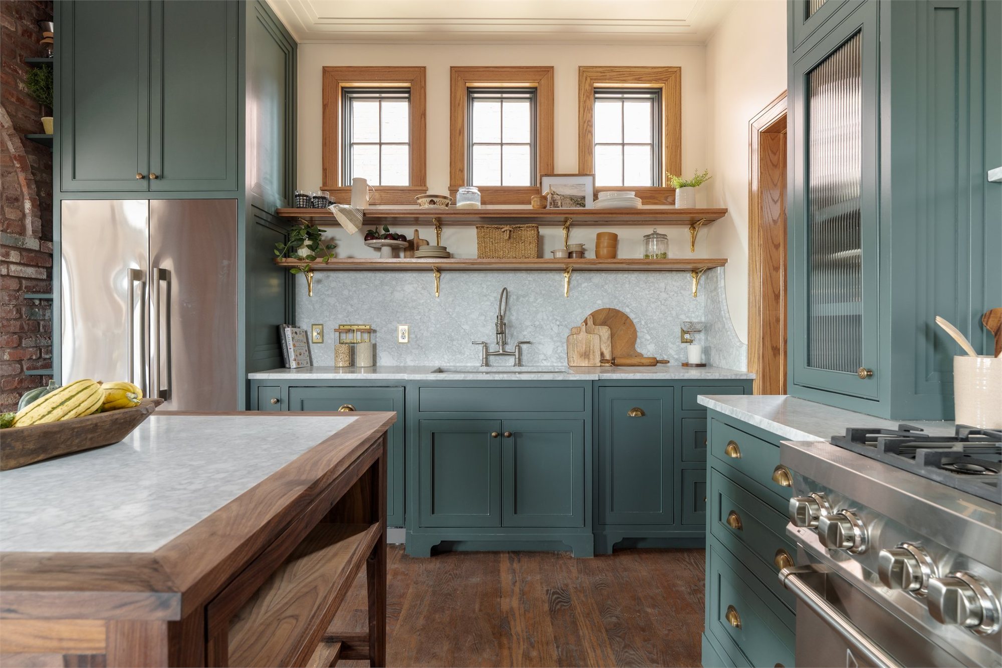 Kitchen with deep teal cabinetry, marble counters, brass cup pulls, and a walnut-trimmed island under three oak-cased windows.