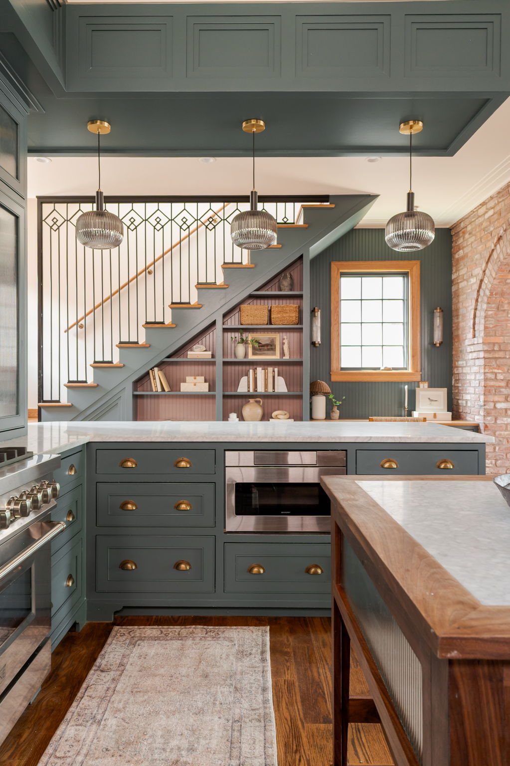 Kitchen framed by an iron-baluster stair — teal cabinetry with brass pulls, marble counters, and a walnut-and-marble island.