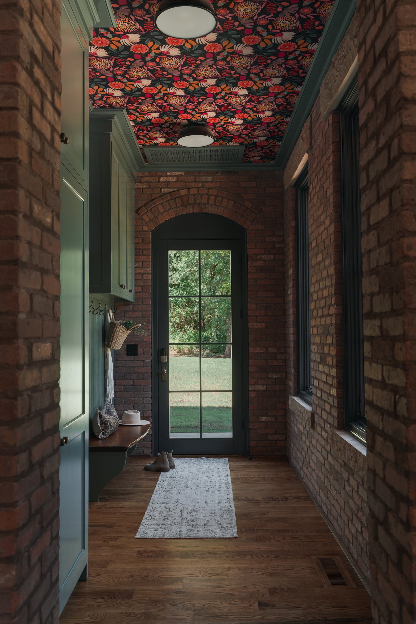 Mudroom corridor — exposed red-brick walls, deep-green millwork, and a floral-papered ceiling above the brick threshold.