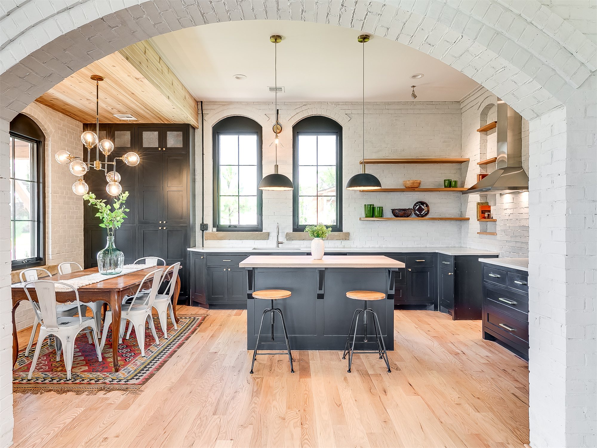 Whitewashed brick arched threshold opening onto a black-cabinet kitchen with butcher-block island and globe-cluster pendants.