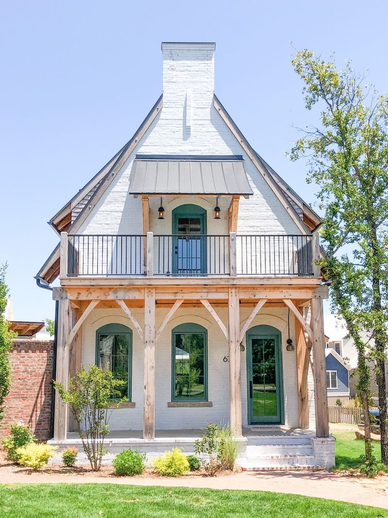 Painted-brick cottage with a peaked gable, timber-frame porch and balcony, and an arched brick threshold in the wall behind.