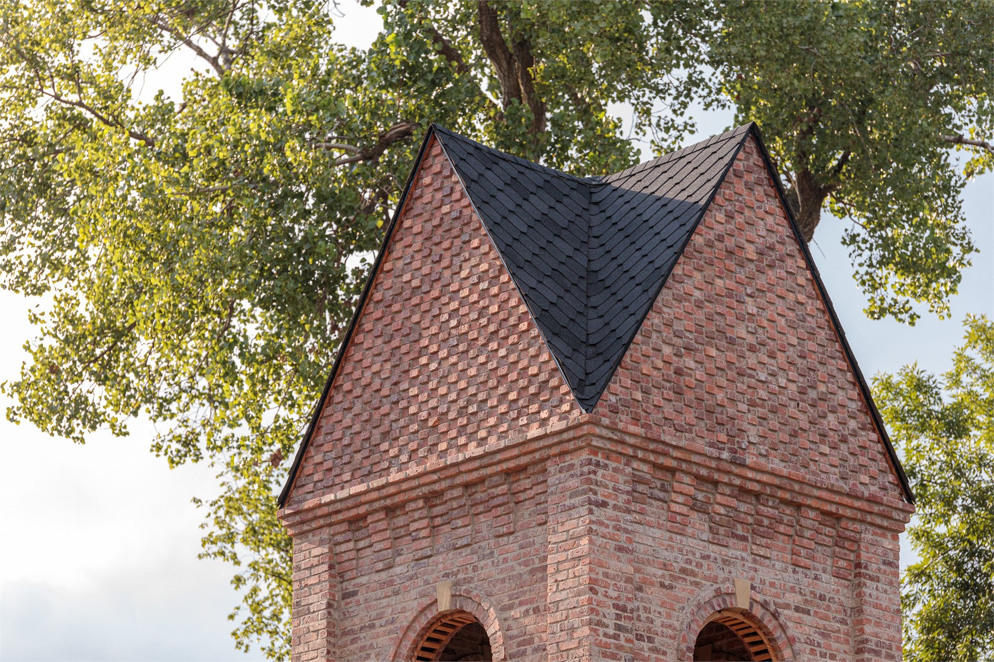 A Selah masonry tower roof — pyramidal slate roof with a single dark-tile face above brick eaves, set in summer foliage.