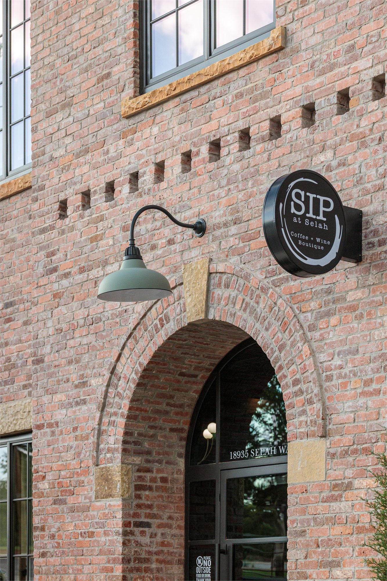 Storefront at Selah — brick facade with sandstone trim, a black gooseneck lamp, and the round 'SIP at Selah' coffee and wine boutique sign.