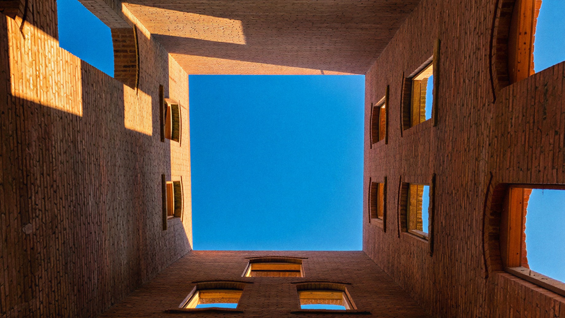 Looking up through a brick tower under construction — blue sky framed by structural masonry walls, golden afternoon light catching the courses.