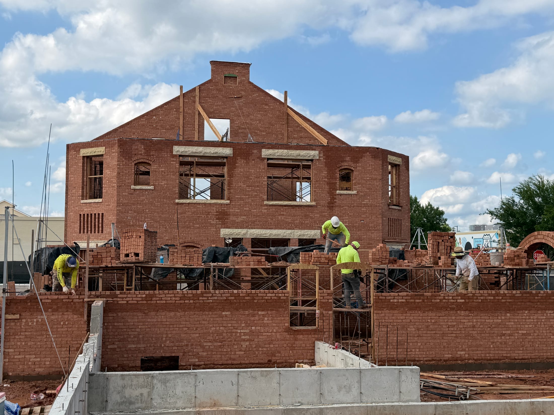 Masons laying structural brick at Townsend in downtown Edmond — a gabled brick building rising behind the courtyard wall under a blue sky.